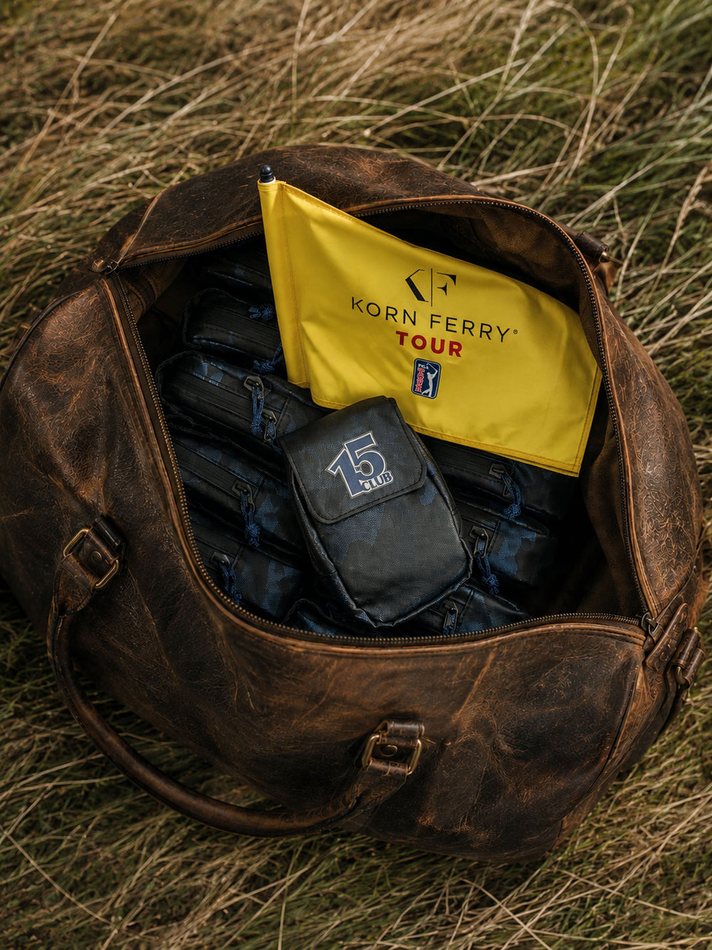 A brown leather duffel bag lies open on dry grass, revealing a bright yellow Korn Ferry Tour flag and several navy camouflage rangefinder cases inside.
